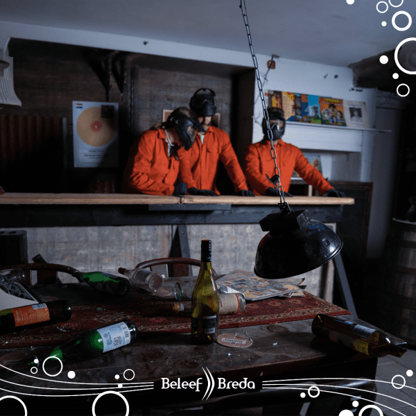 drie mannen in oranje overals die in de smash room van BeleefBreda staan. Ook liggen er verschillende flessen en lampen.